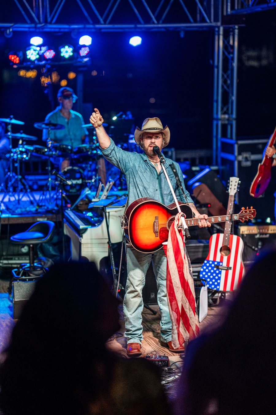 Joshua Alan pointing to the crowd under stage lights, drummer and flag guitar behind him