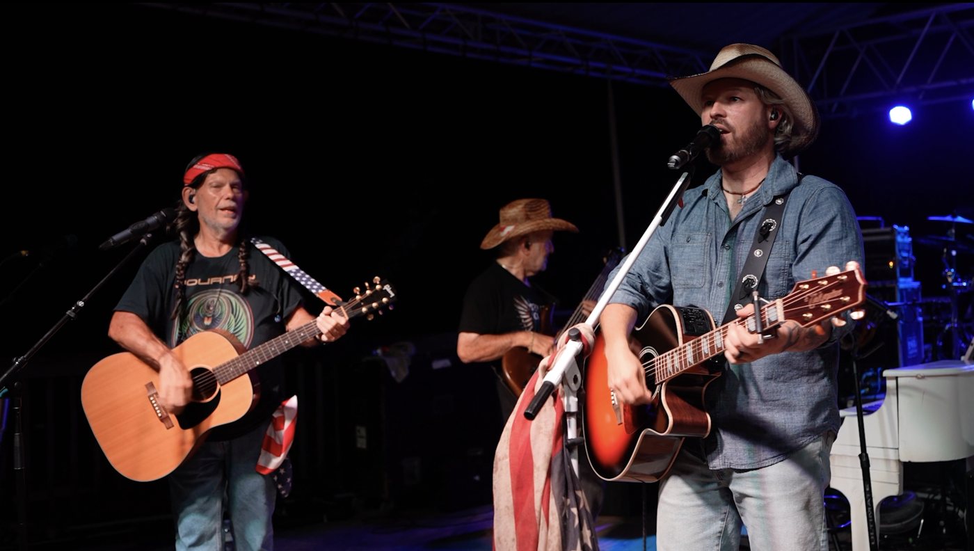 Rod Burton as Willie Nelson, in a red bandana and braids, playing acoustic guitar beside Joshua “Toby” Alan on stage